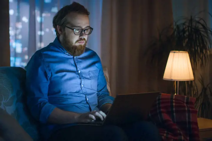 Man med blå skjorta, skägg och glasögon sitter med laptop i soffan och lampan intill tänd