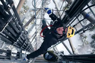 En man med gul hjälm klättar högt upp i en mast. Bilden ses ovanifrån och mannen tittar upp mot kameran.