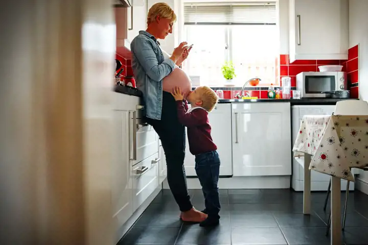 En gravid kvinna står i ett kök och ett litet barn pussar på magen